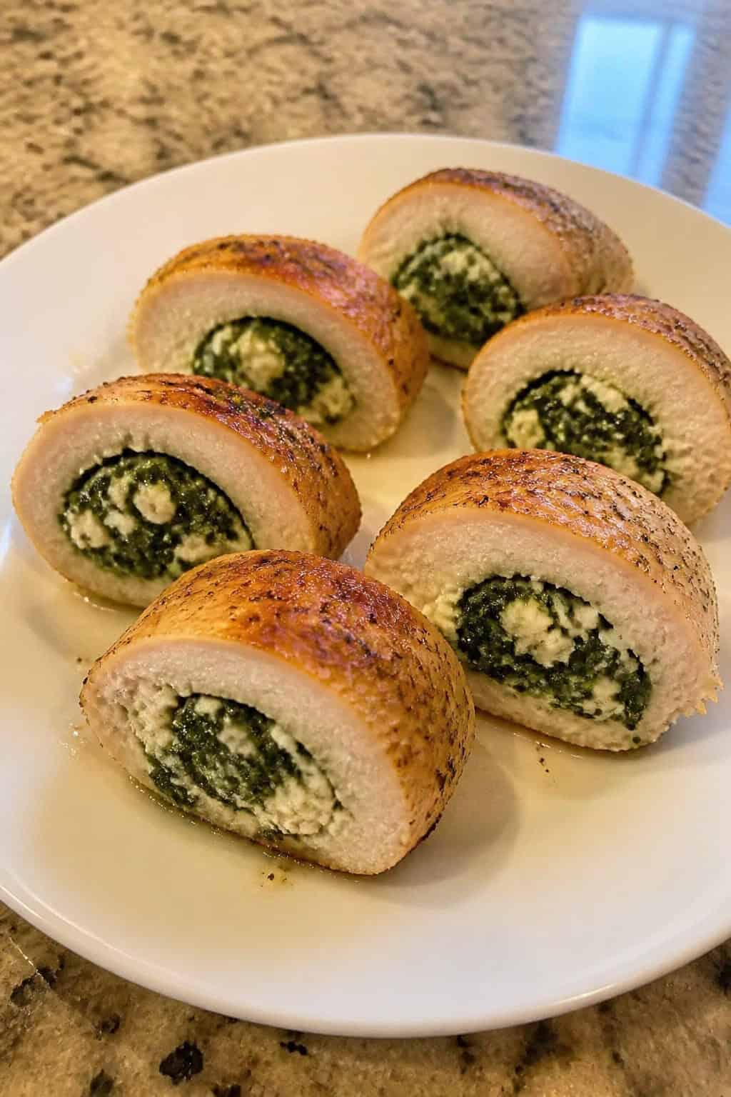 Close-up of sliced spinach and feta stuffed chicken rounds on a wooden board with spinach leaves and feta cheese garnishes.