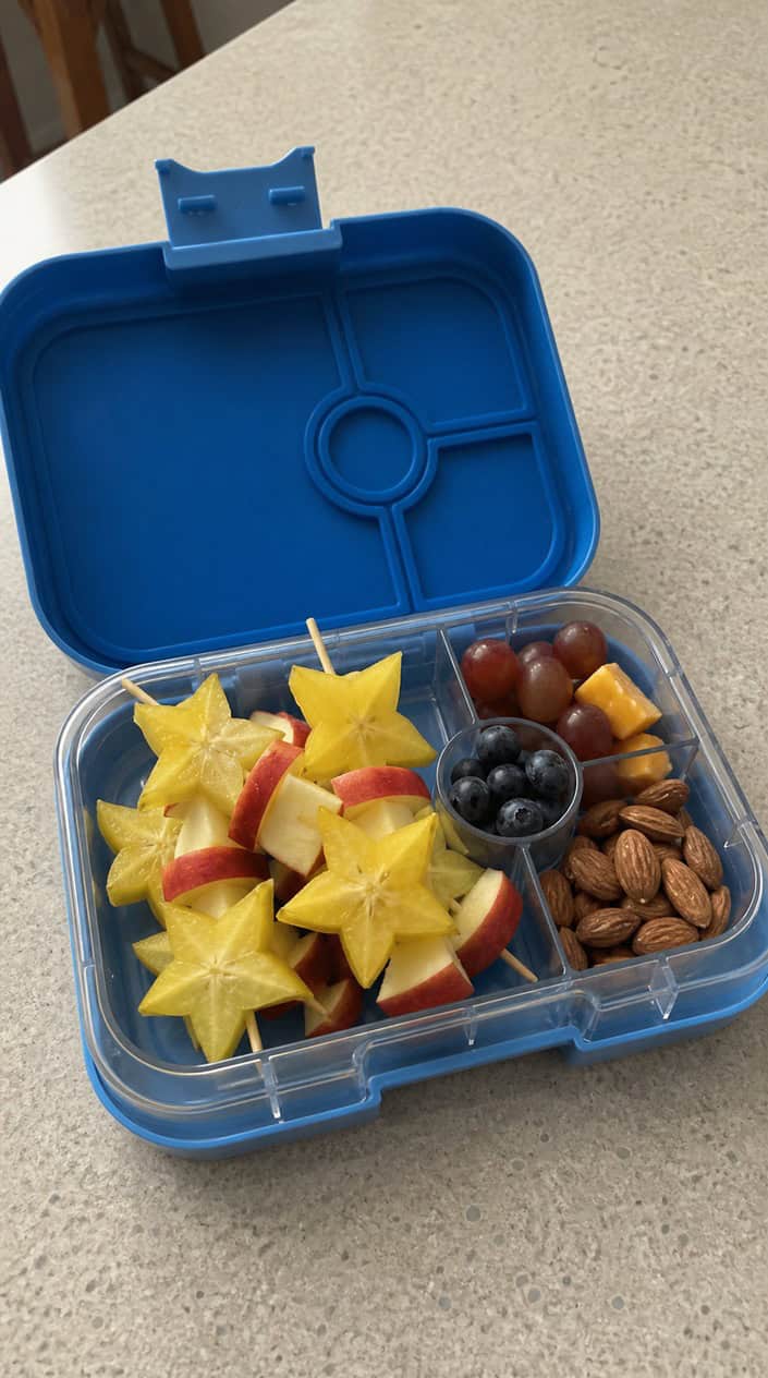 An open lunchbox on a wood table showing four short skewers alternating star-shaped starfruit slices and pale yellow apple chunks.