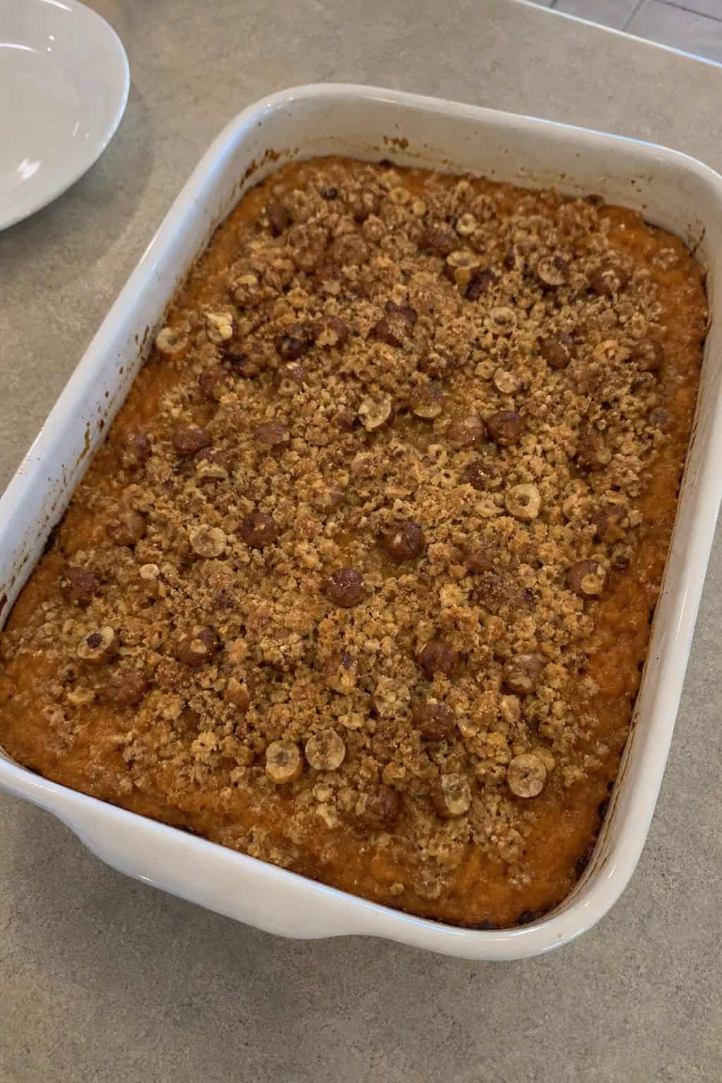 A baked sweet potato casserole topped with brown butter and toasted hazelnuts on a wooden table with autumn ingredients around it.