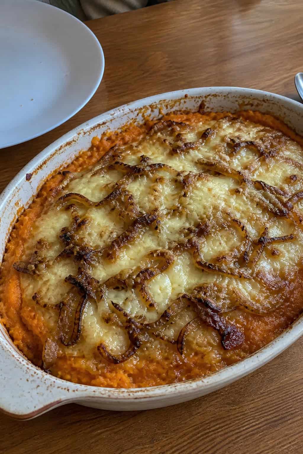 A baked sweet potato casserole topped with caramelized onions and melted cheese in a ceramic dish on a wooden table with fresh ingredients around it.