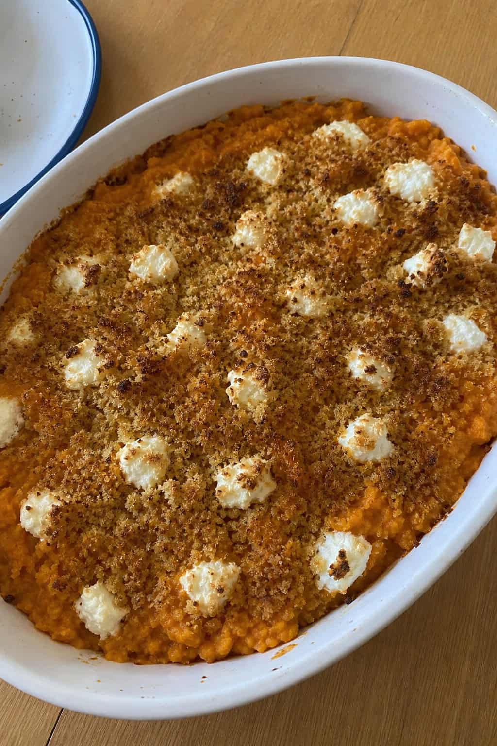 A sweet potato casserole topped with goat cheese and thyme in a ceramic dish on a wooden table with fresh thyme and whole sweet potatoes nearby.