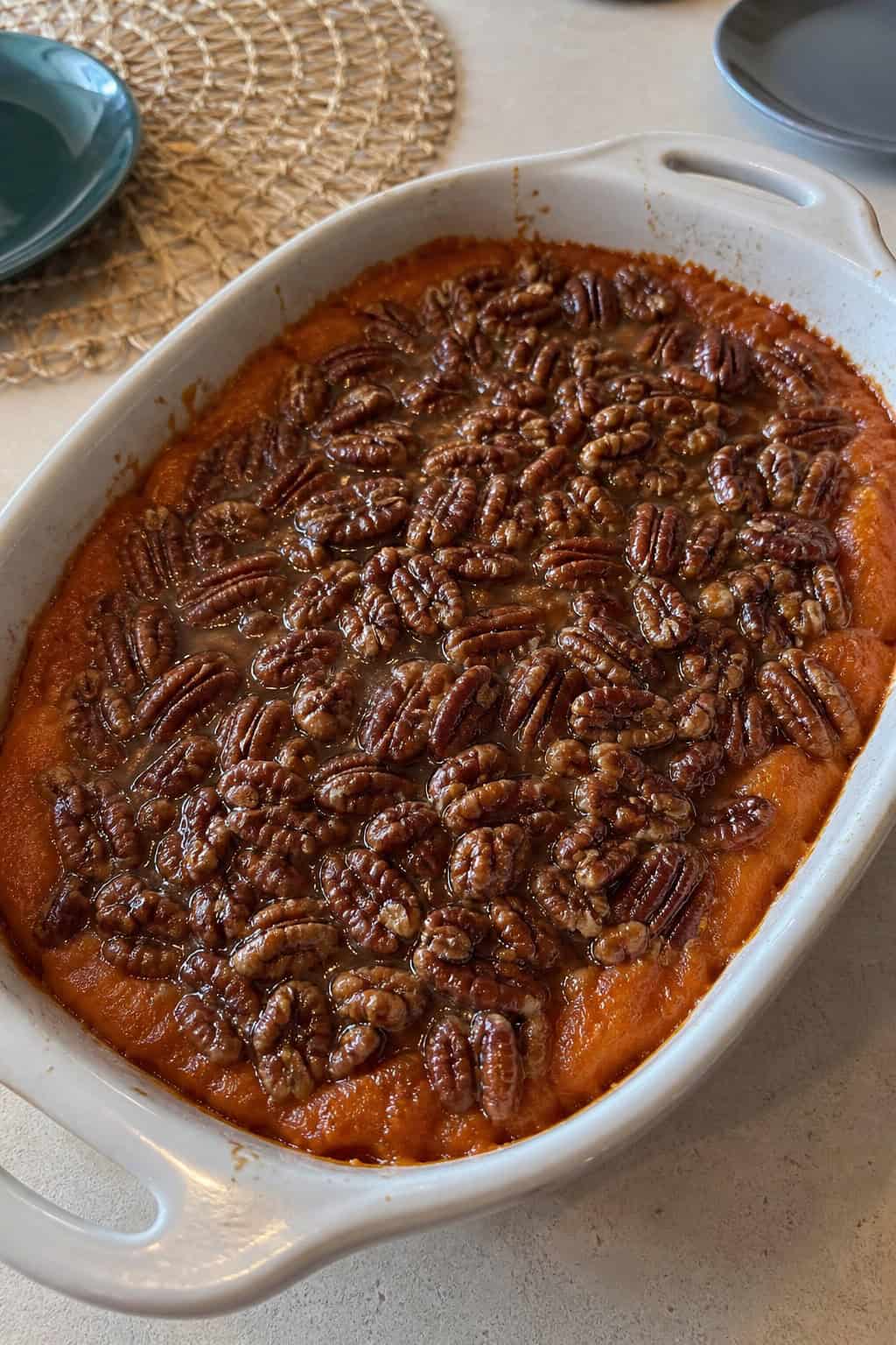 A baked sweet potato casserole topped with glazed pecans in a ceramic dish on a wooden table with autumn decorations.