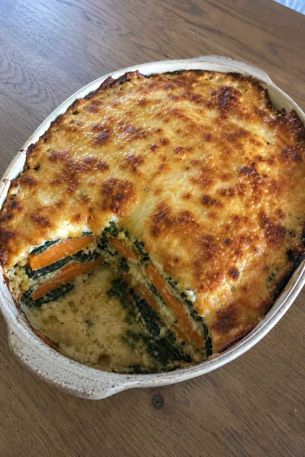 A baked sweet potato lasagna casserole with spinach in a ceramic dish on a wooden table.