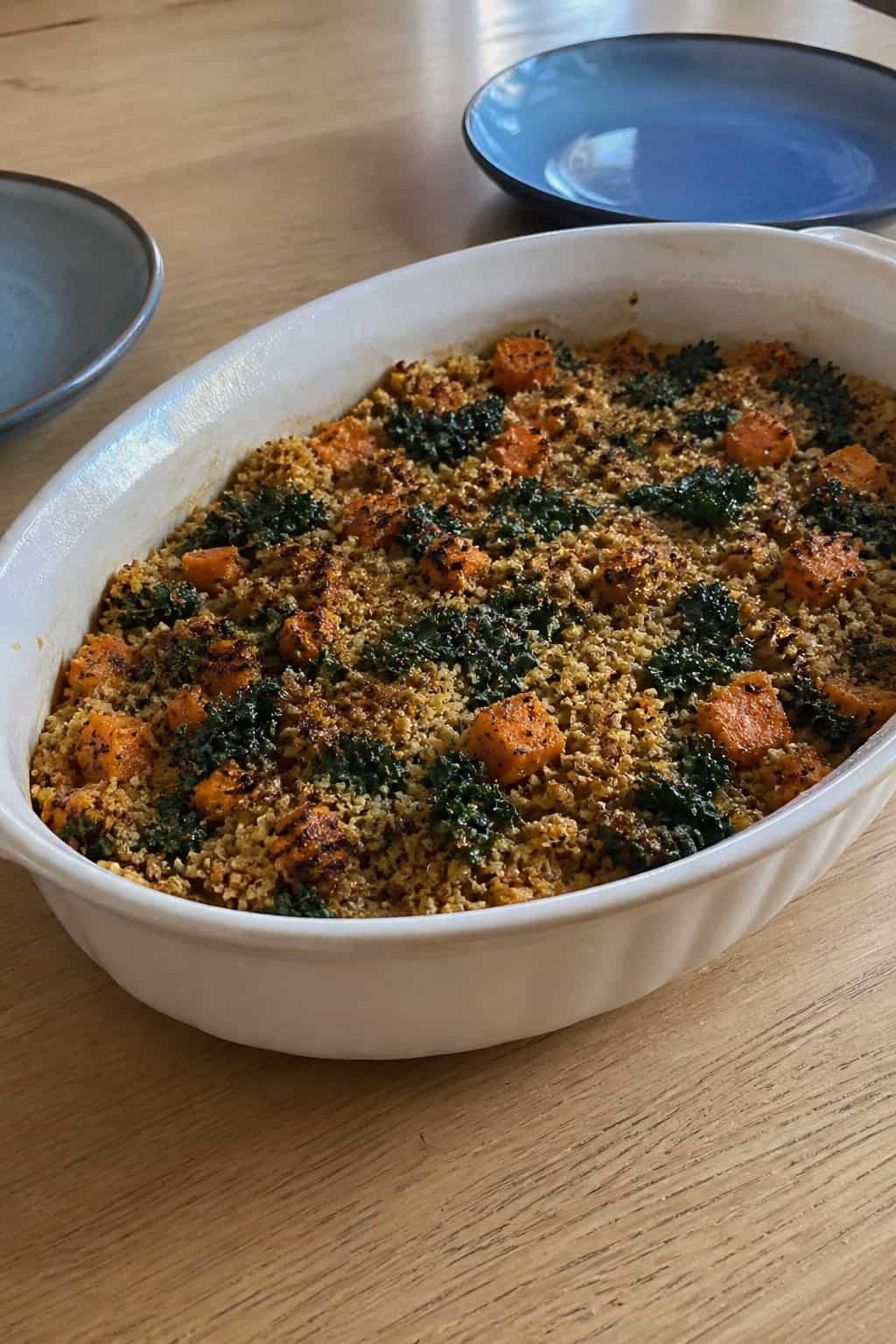 A baked casserole dish filled with sweet potato, quinoa, and kale on a wooden table with fresh ingredients around it.