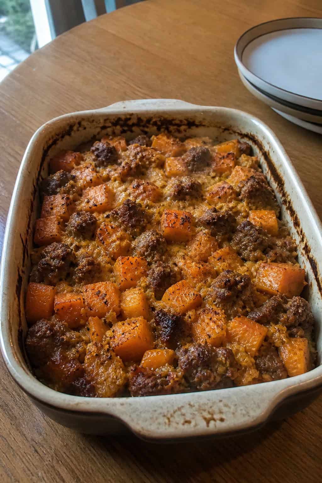 A freshly baked sweet potato, sausage, and sage casserole in a rustic baking dish on a wooden table.