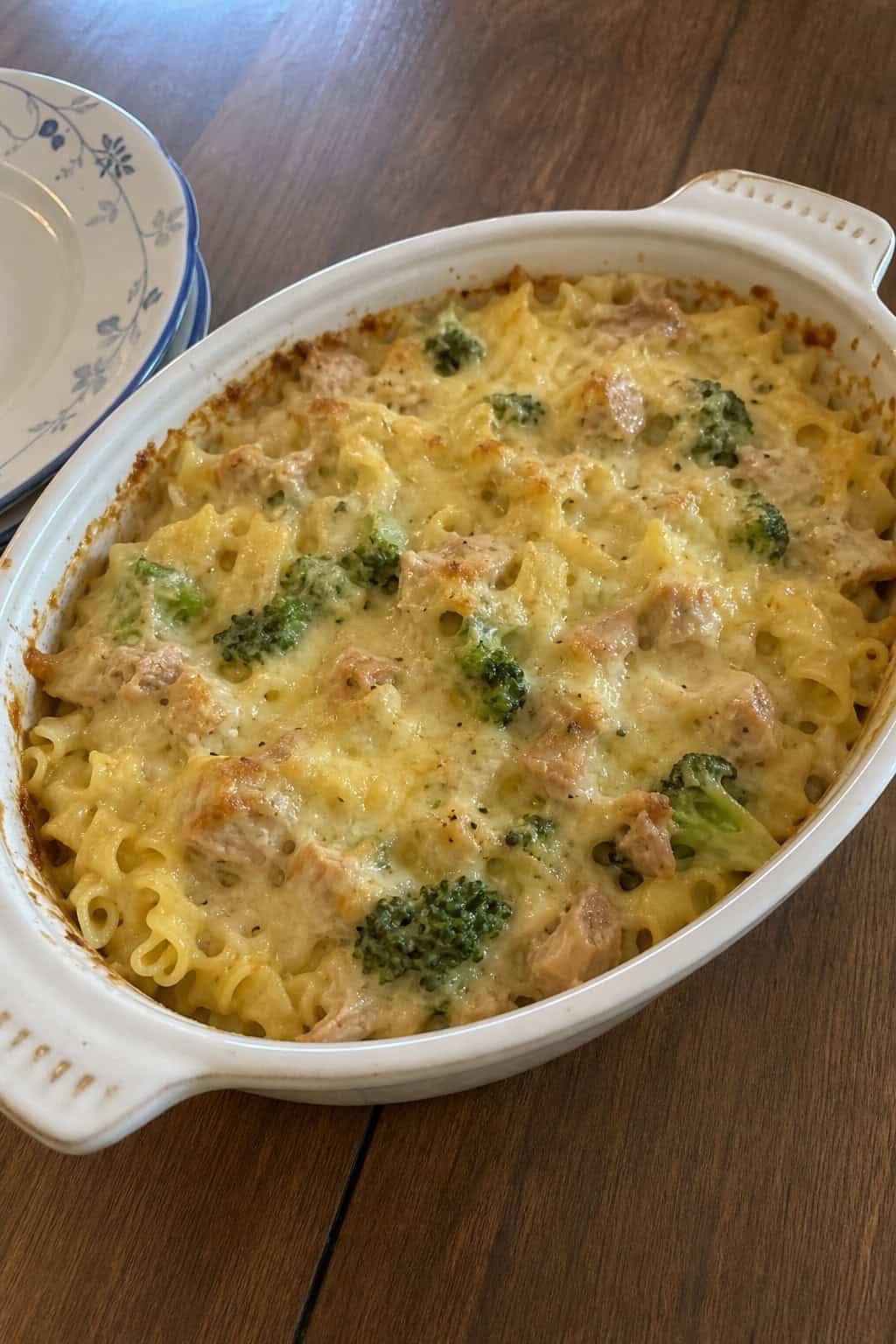 A baked tuna noodle casserole with broccoli and melted white cheddar cheese in a ceramic dish on a wooden table.