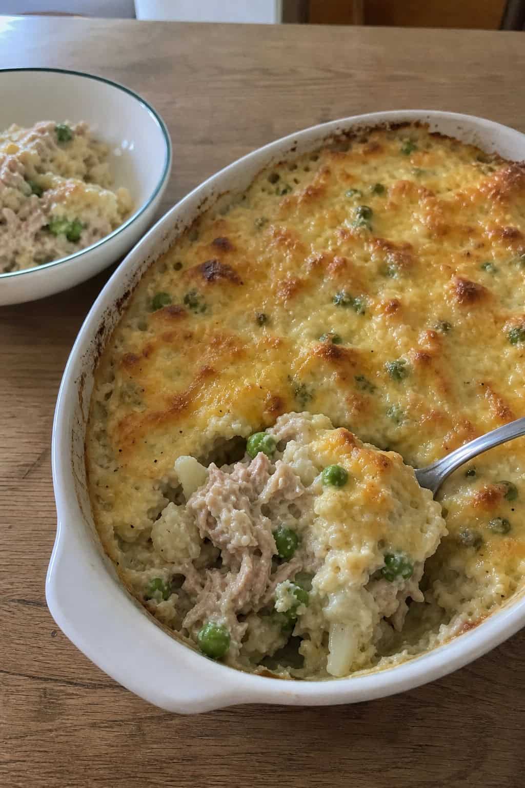 A tuna noodle casserole made with cauliflower rice in a ceramic baking dish on a wooden table, with fresh herbs and lemon nearby.