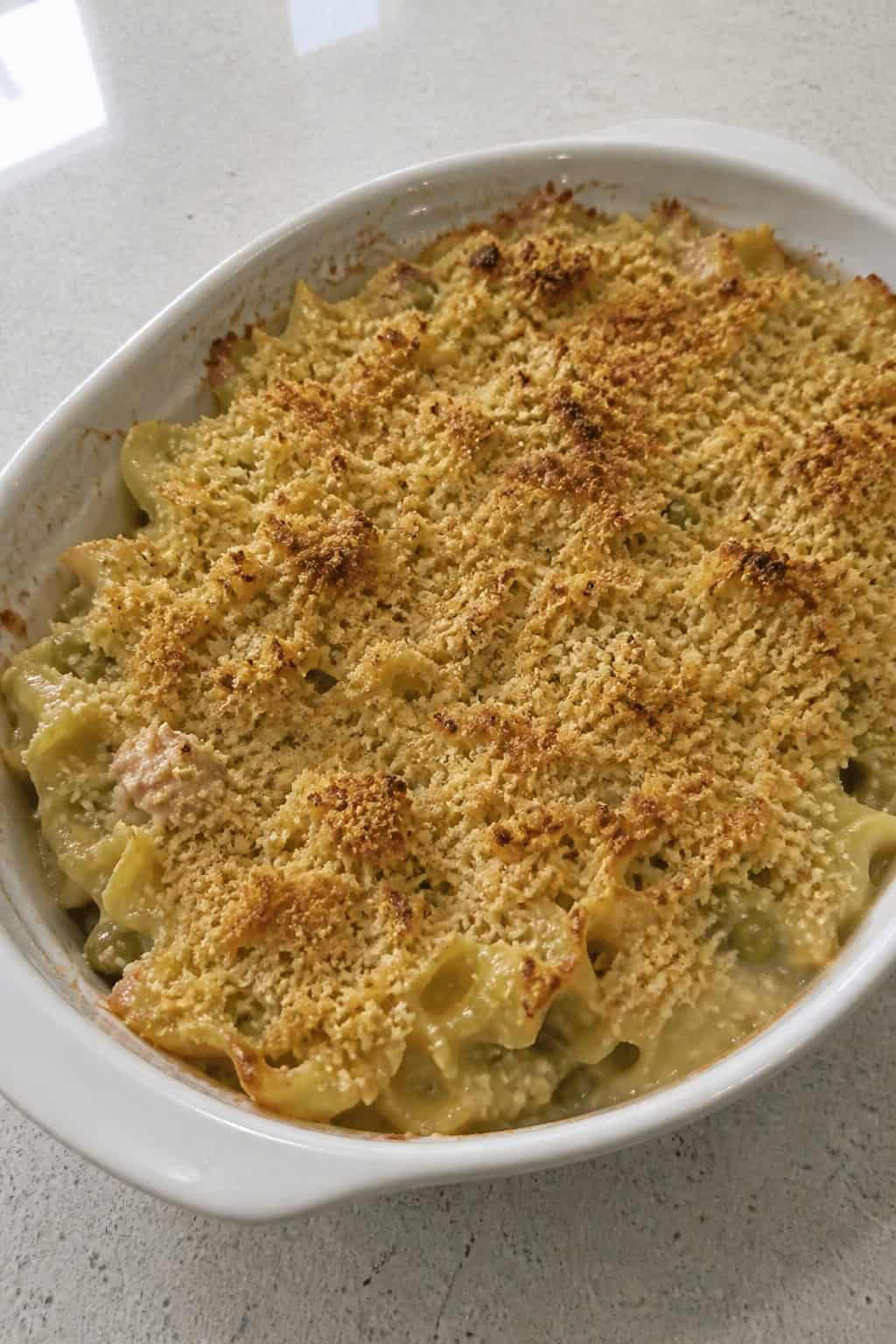 A baked tuna noodle casserole in a ceramic dish garnished with fresh parsley on a wooden table with small bowls of spices nearby.
