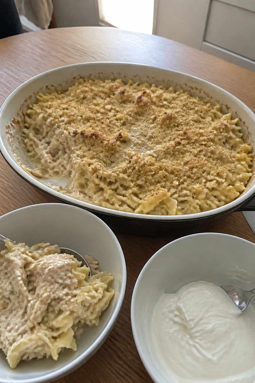 A baked tuna noodle casserole in a ceramic dish on a wooden table with a bowl of Greek yogurt sauce and fresh herbs nearby.