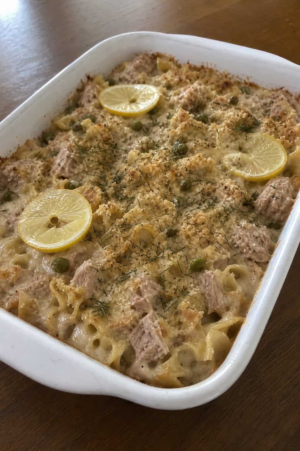 A baked tuna noodle casserole garnished with lemon slices, dill, and capers on a wooden table.