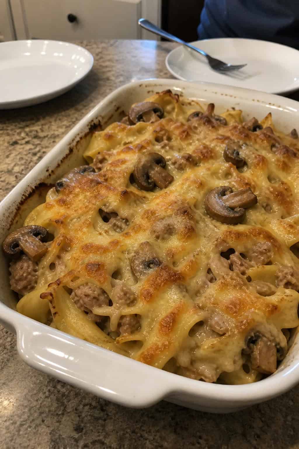 A baked tuna noodle casserole with melted Swiss cheese and mushrooms in a ceramic dish on a wooden table with fresh ingredients nearby.