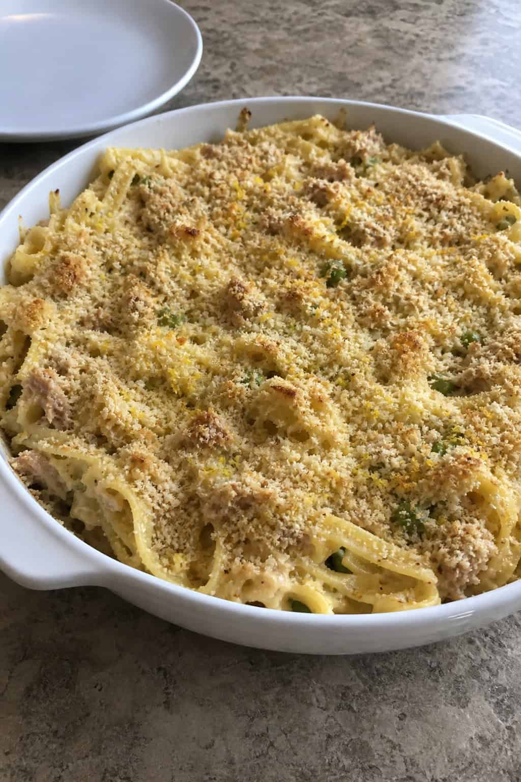 A baked tuna noodle casserole topped with melted cheese and lemon zest in a ceramic dish on a wooden table with a lemon half and Parmesan cheese nearby.