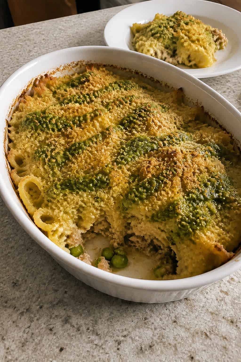 25 Tuna Noodle Casseroles for Busy Weeknights 20 A baked tuna noodle casserole with peas and green pesto in a ceramic dish on a wooden table with fresh ingredients around it.