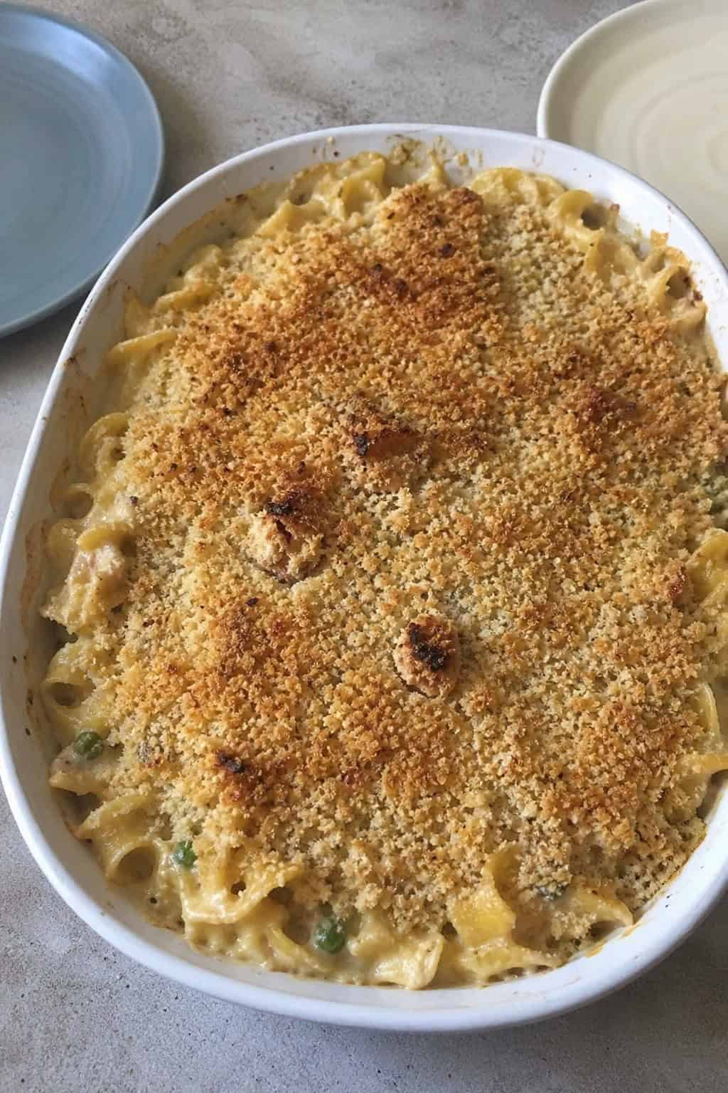 25 Tuna Noodle Casseroles for Busy Weeknights 6 A baked tuna noodle casserole in a ceramic dish garnished with thyme and roasted garlic on a wooden table.