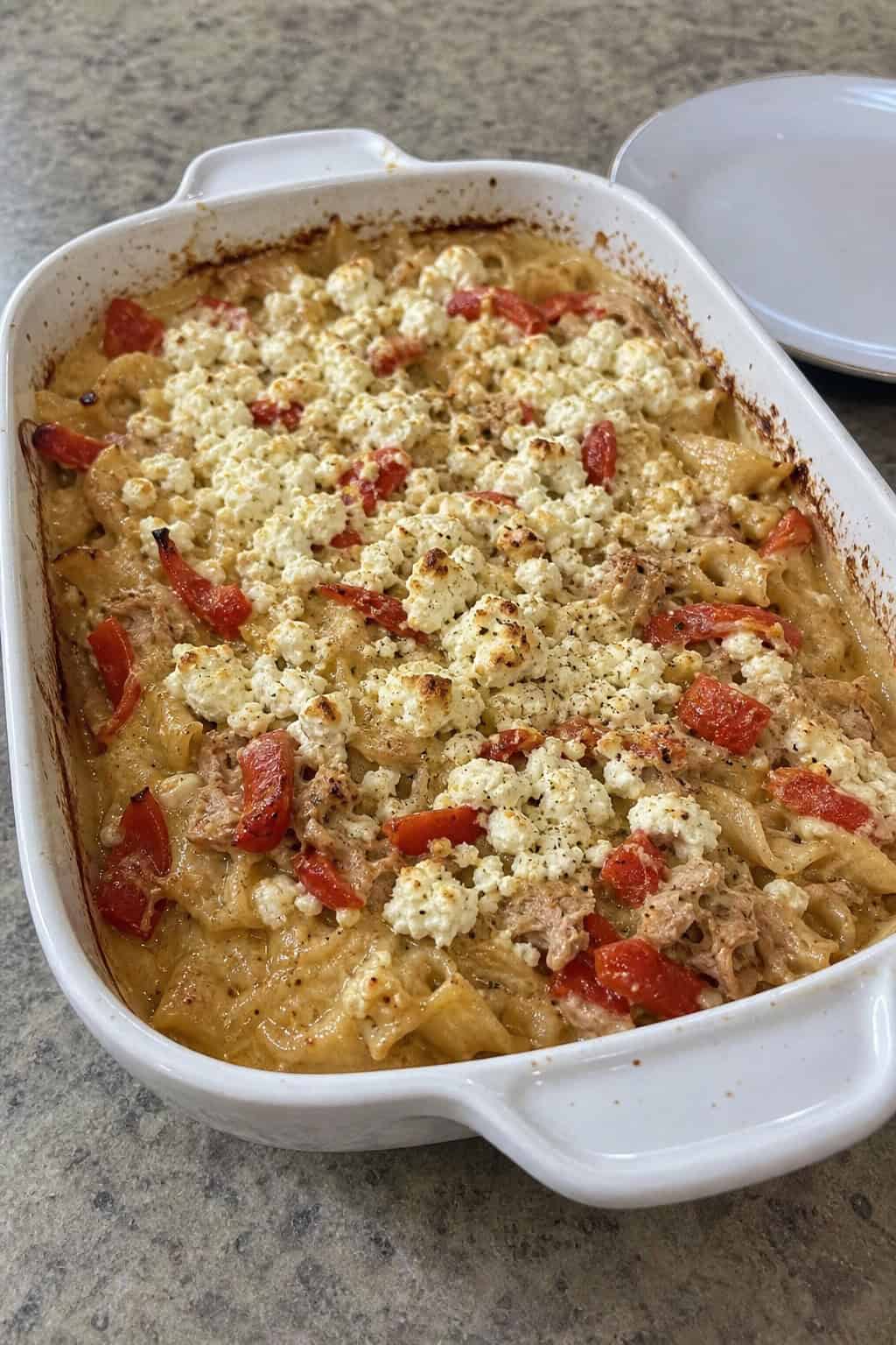 A baked tuna noodle casserole with roasted red peppers and feta cheese in a ceramic dish on a wooden table.