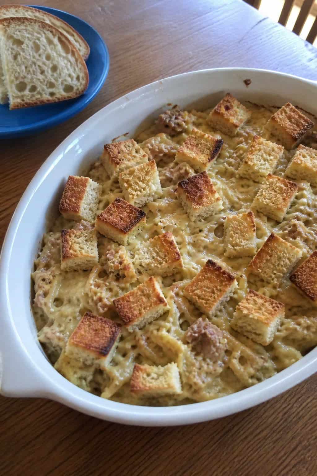 A baked tuna noodle casserole topped with crispy sourdough croutons in a ceramic dish on a wooden table with fresh bread and cheese nearby.