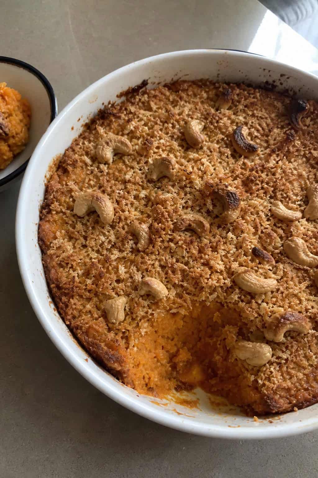 A golden-brown sweet potato casserole topped with cashews and coconut flakes in a ceramic dish on a wooden table with fresh sweet potatoes and cashews nearby.