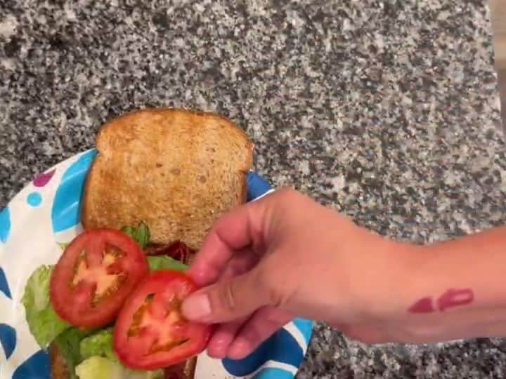 A hand places a tomato slice onto a toasted sandwich already layered with lettuce and bacon on a patterned paper plate.