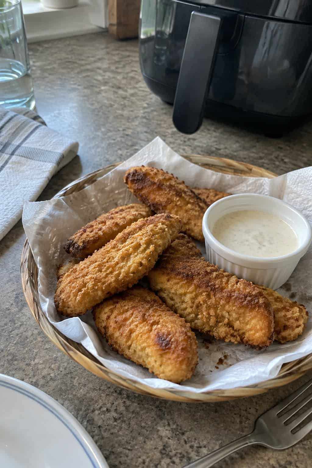 A plate of golden chicken tenders sits beside a small bowl of dipping sauce on a kitchen counter.