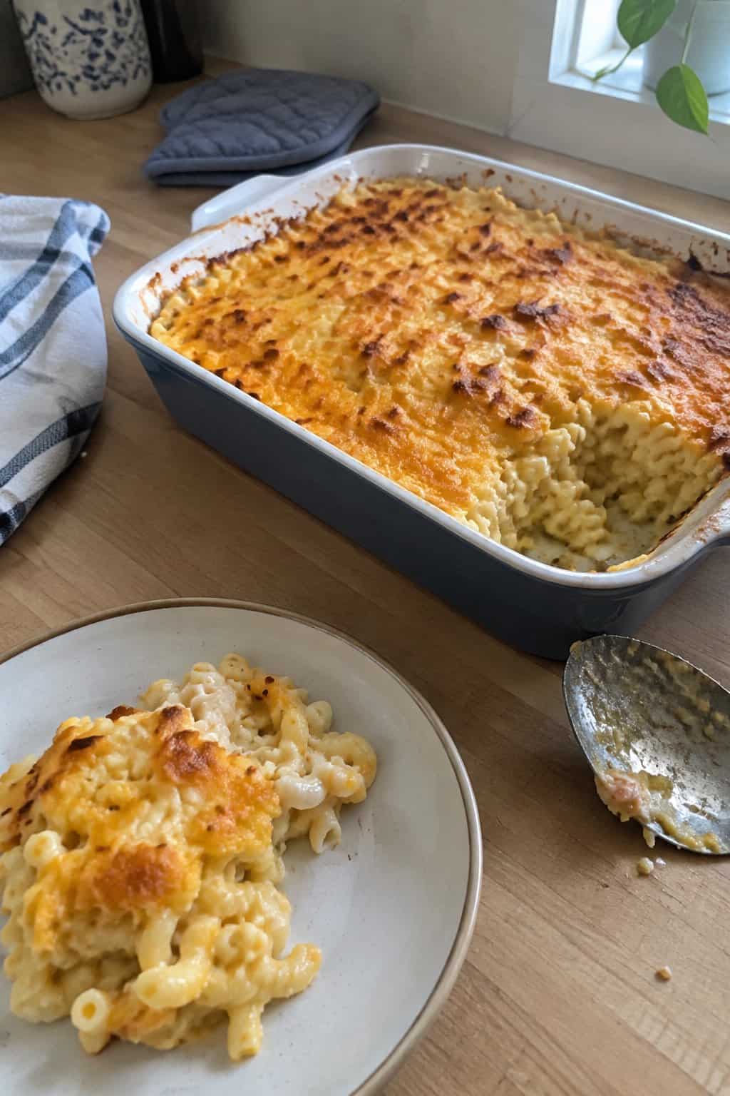A casserole dish of baked macaroni and cheese with a golden browned topping sits on a kitchen counter beside a serving plate.