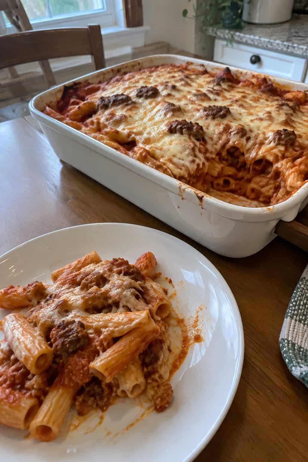 A serving of baked ziti with Italian sausage and melted cheese beside a casserole dish.