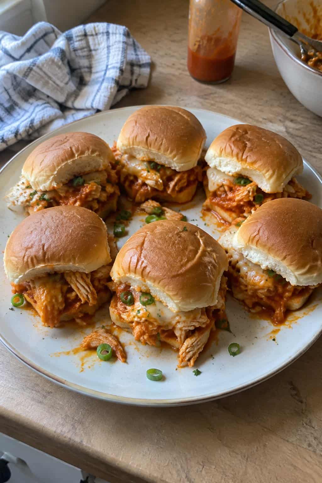 A plate of homemade Buffalo chicken sliders with shredded chicken, melted cheese, and green onion on a kitchen table.
