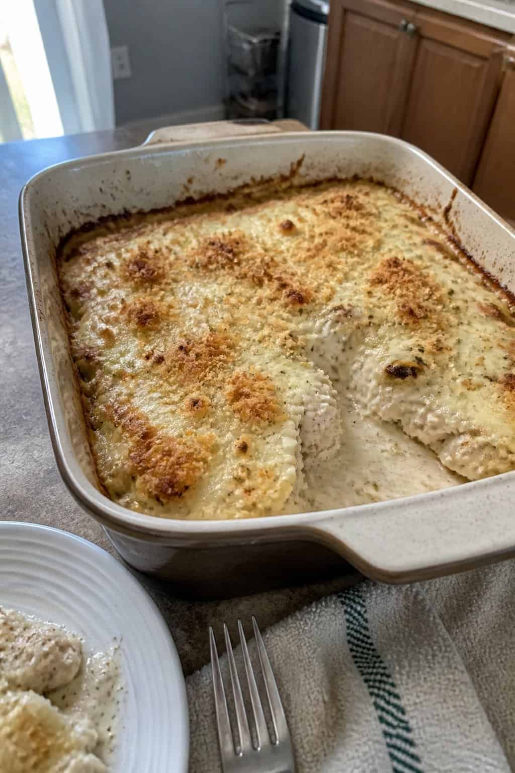 A partially served creamy chicken Parmesan casserole in a ceramic baking dish on a kitchen table.