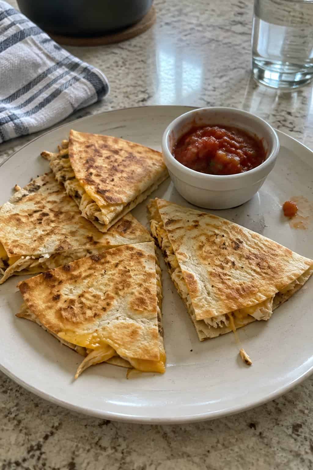 A plate of homemade chicken quesadilla wedges with melted cheese and a bowl of salsa on a kitchen table.
