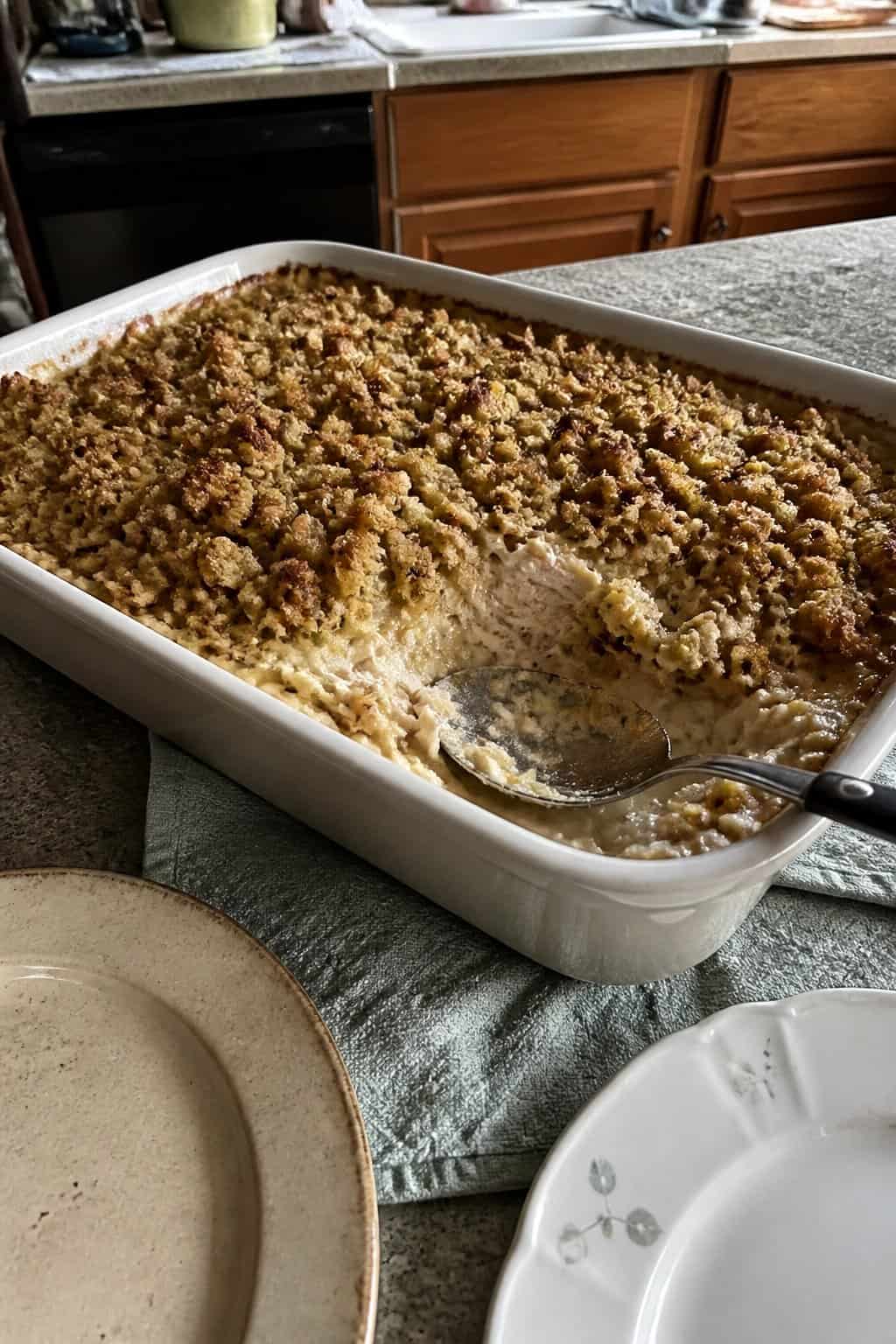 A baking dish of creamy chicken and stuffing casserole sits on a kitchen table beside everyday plates and a serving spoon.