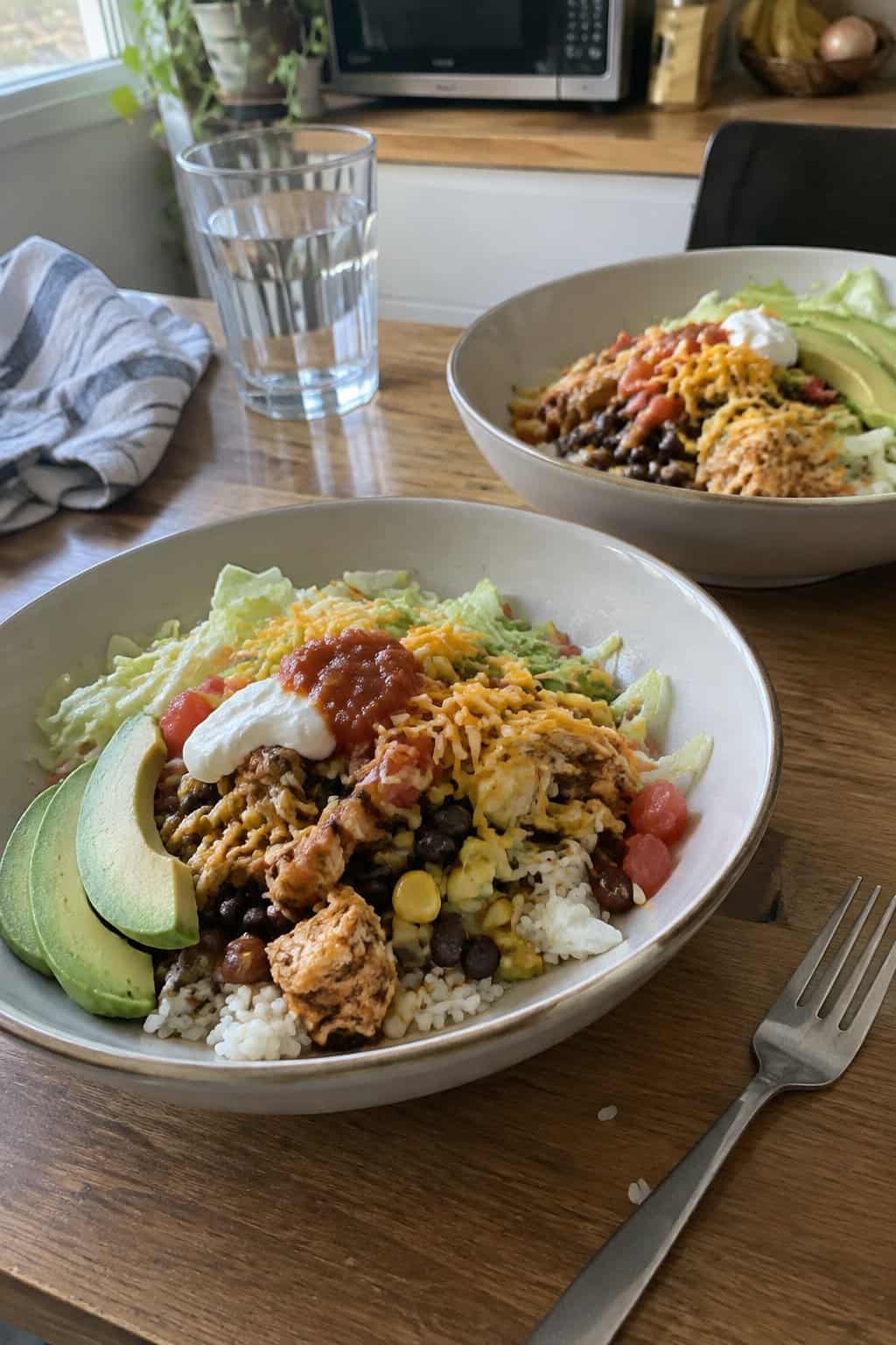 Two bowls of chicken taco rice topped with beans, corn, vegetables, cheese, and avocado on a kitchen table.