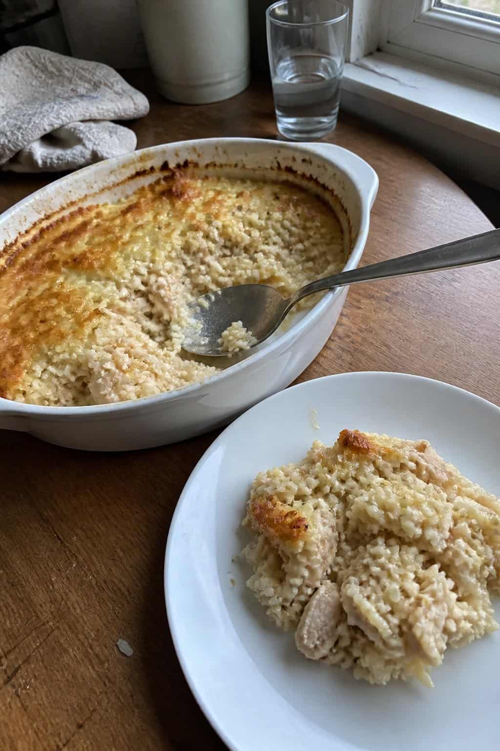 A homemade chicken and rice casserole in a ceramic baking dish beside a serving of casserole on a plate.