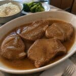 Tender boneless pork chops covered in brown gravy in a serving dish beside simple dinner sides.