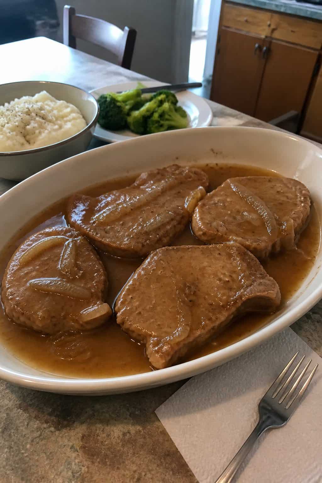 Tender boneless pork chops covered in brown gravy in a serving dish beside simple dinner sides.