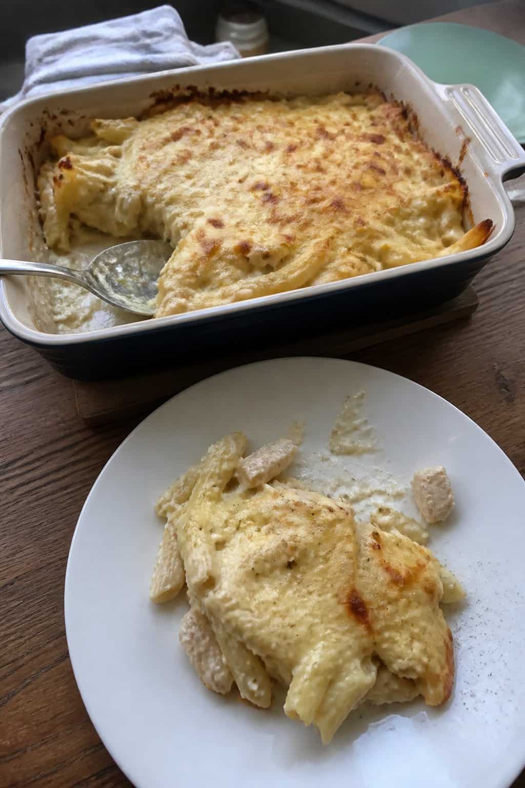 A creamy chicken Alfredo pasta bake in a ceramic dish with a serving of pasta on a plate beside it.