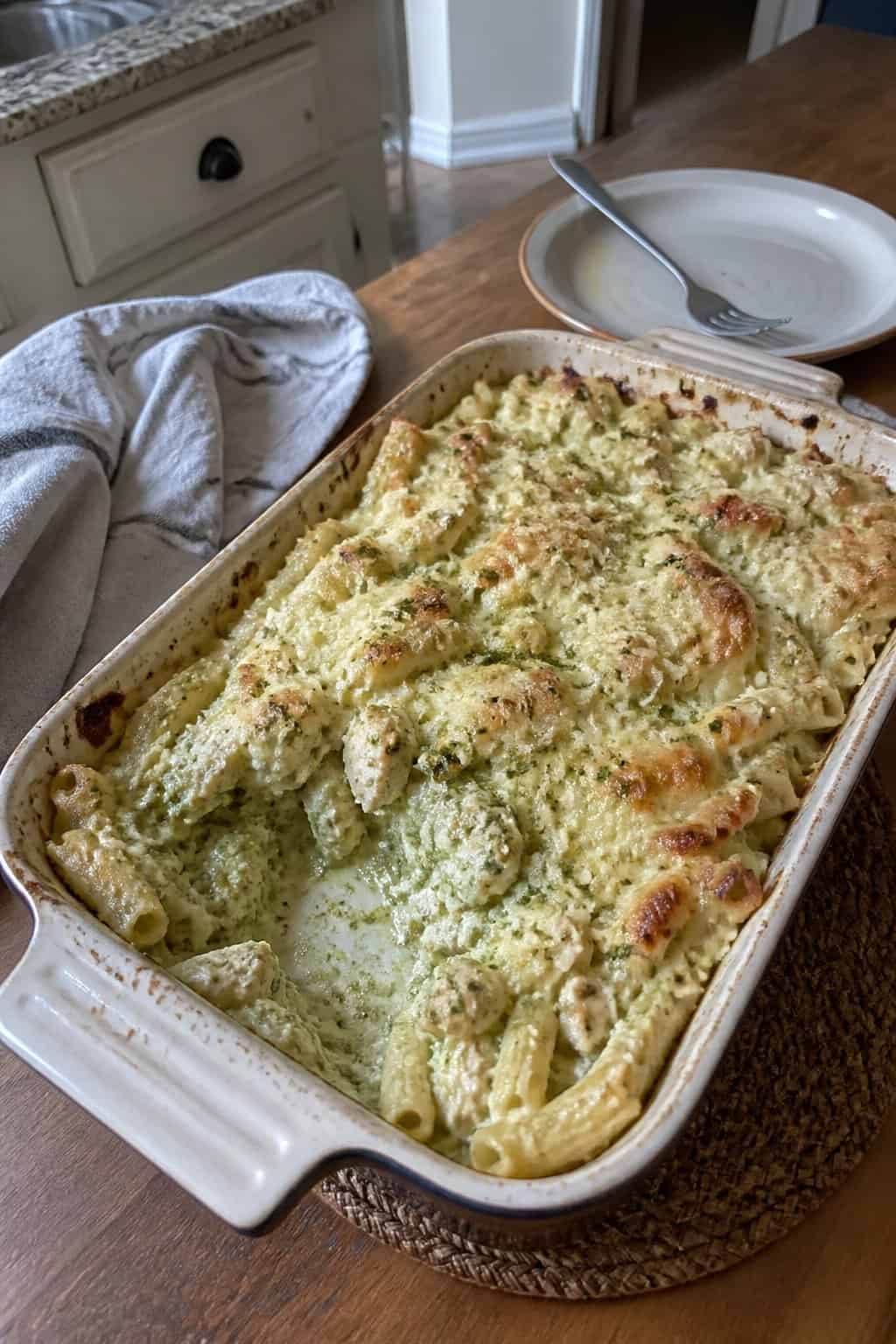 A baked pasta casserole with chicken, creamy pesto sauce, and melted cheese in a ceramic dish on a kitchen table.