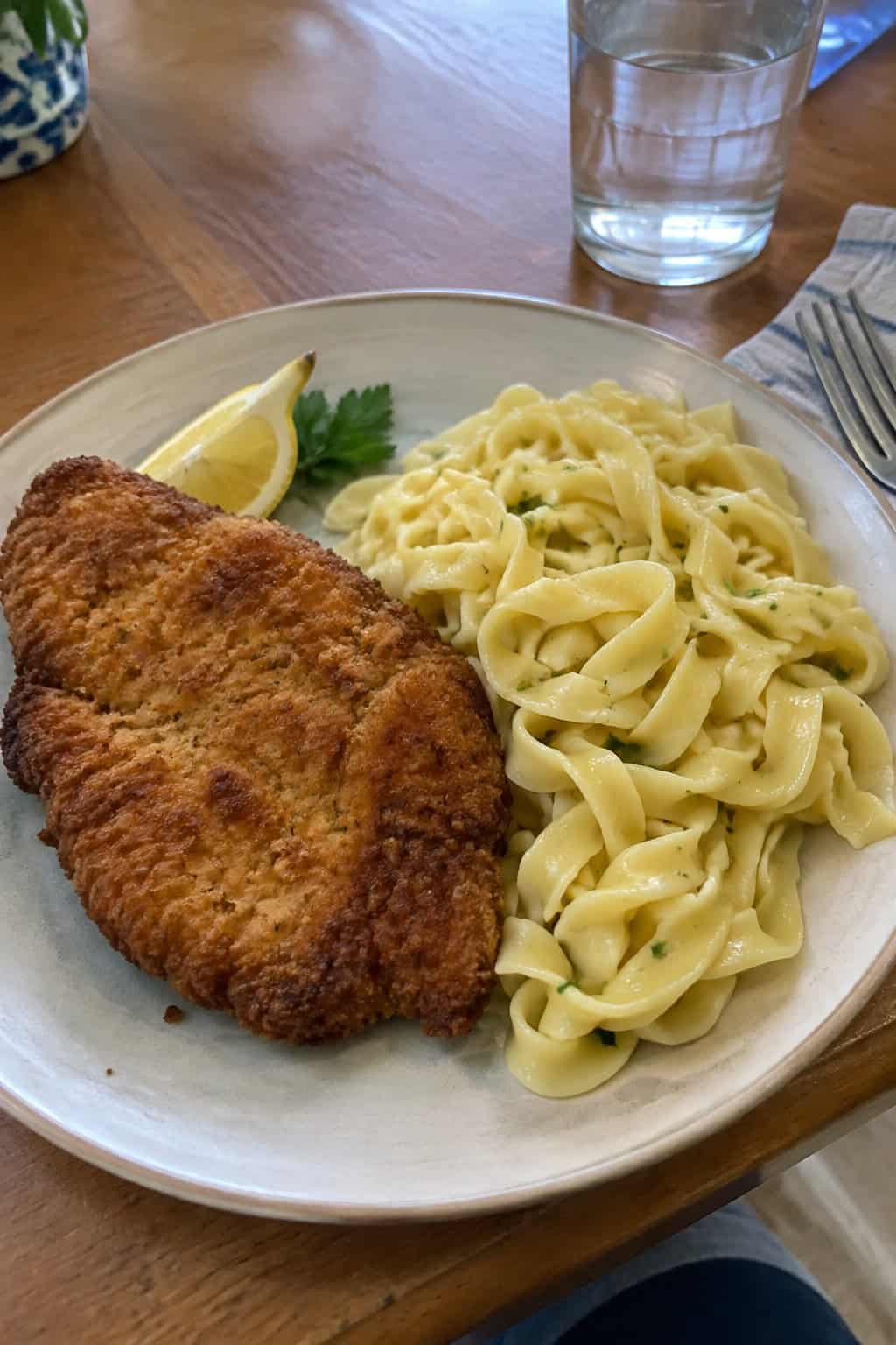 A breaded chicken cutlet served with buttered noodles and a lemon wedge.