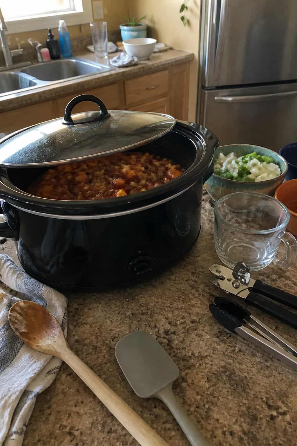 A black slow cooker sits on a kitchen counter beside cooking utensils, bowls, and prepared vegetables.