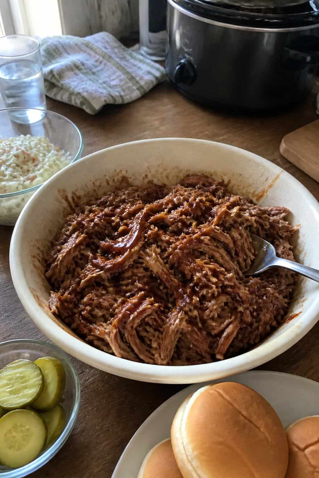 A bowl of barbecue pulled pork sits on a kitchen table beside simple side dishes.