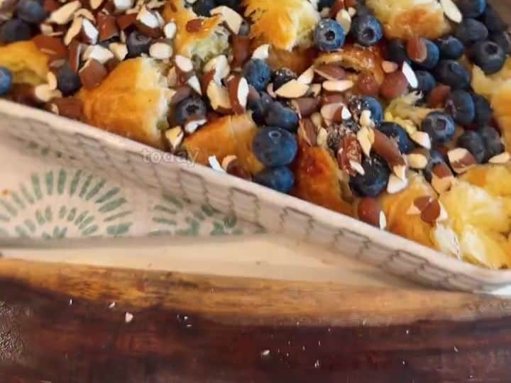 Blueberry Croissant Bake 4 A hand sprinkles chopped almonds over torn croissant pieces topped with blueberries in a baking dish.