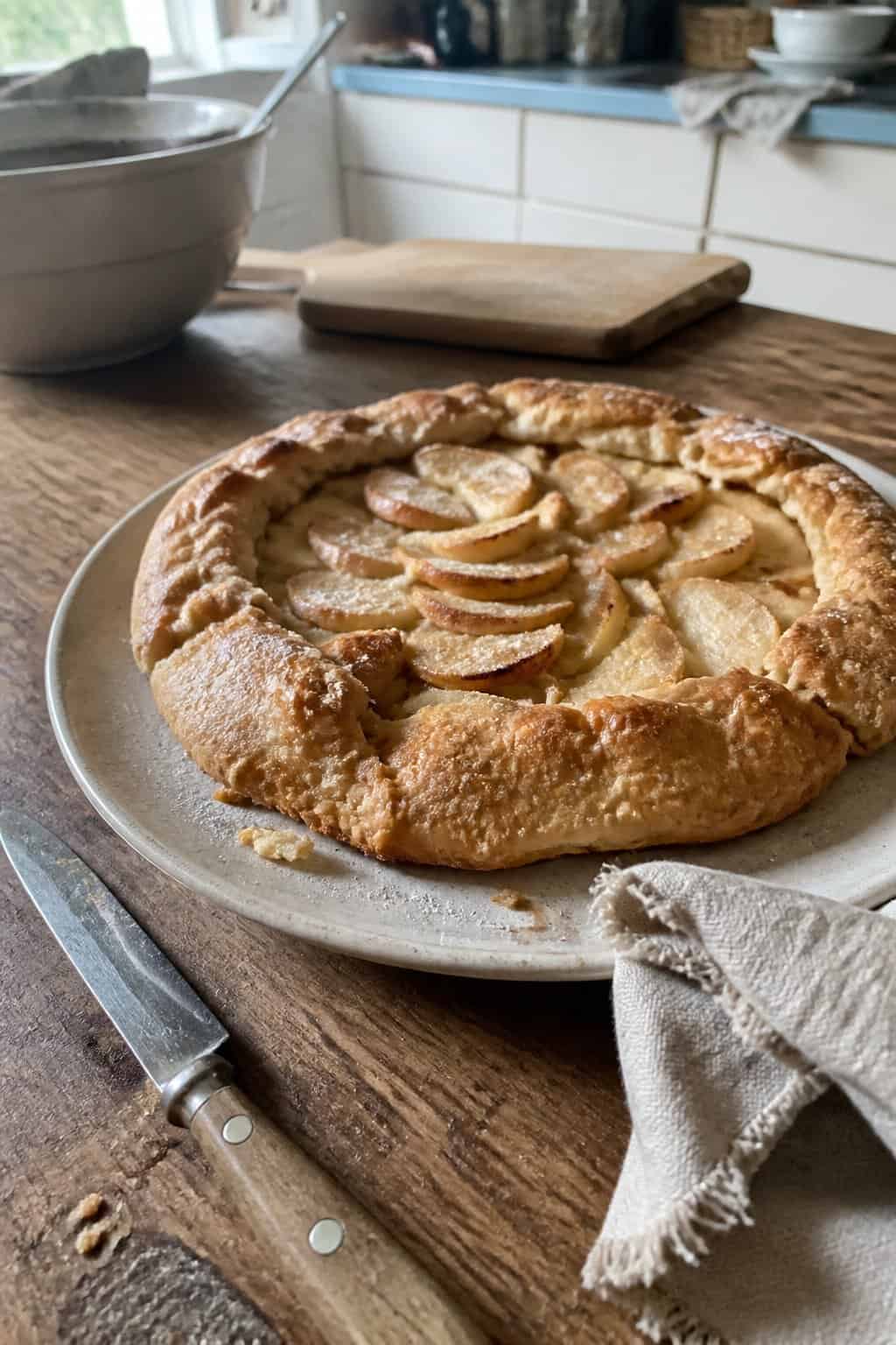A homemade pear and apple galette sits on a plate on a kitchen table beside a knife and napkin.