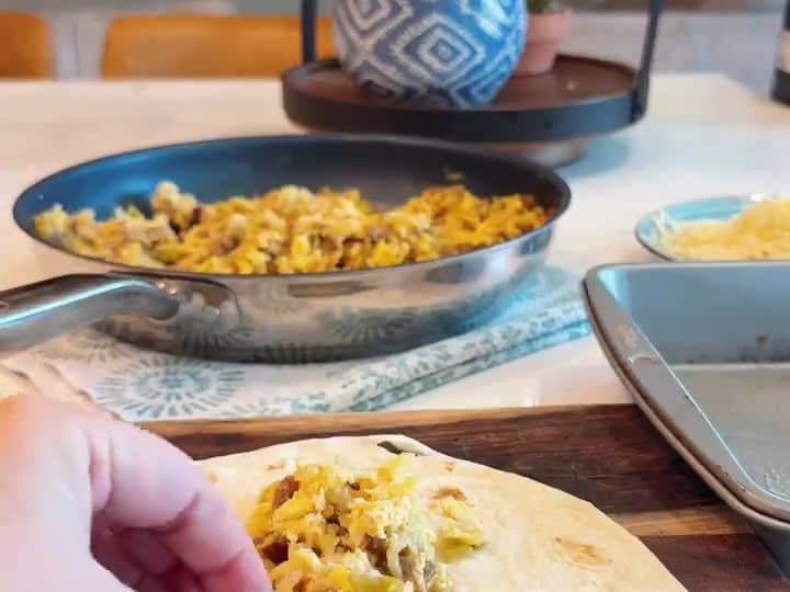 A hand places seasoned sausage, egg and hash brown filling onto an open flour tortilla ready to be rolled.