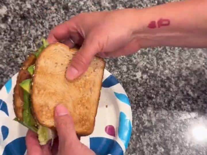 A hand presses down the final toasted bread slice onto the completed BLTA sandwich on a paper plate.