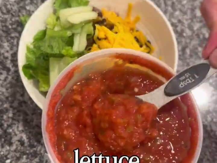 A finished taco bowl topped with lettuce, shredded cheese, and a scoop of red salsa being added with a spoon, held by a hand.