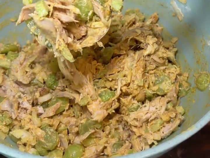 A large bowl of finished curried chicken salad, evenly coated in dressing with visible chicken and grapes, being stirred with a spoon.