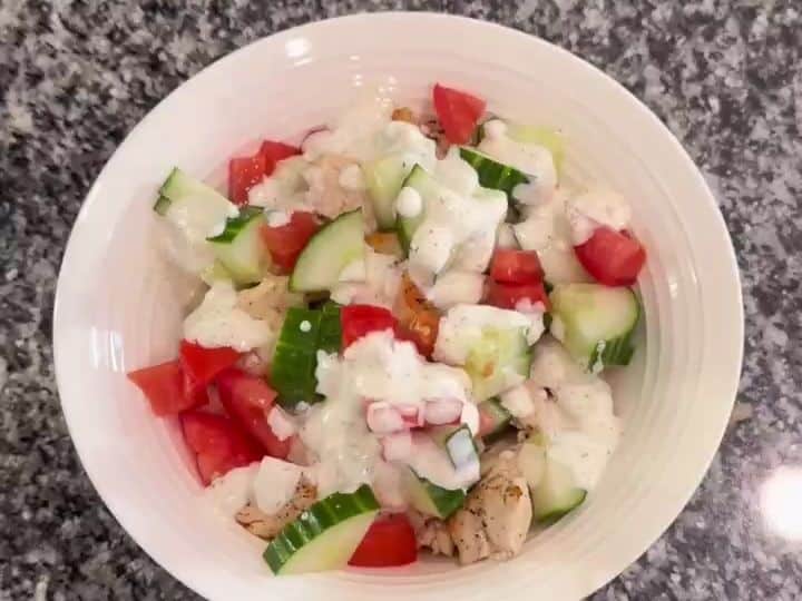 A completed Greek bowl with rice, tomatoes, cucumbers, chicken, and crumbled feta cheese on top.