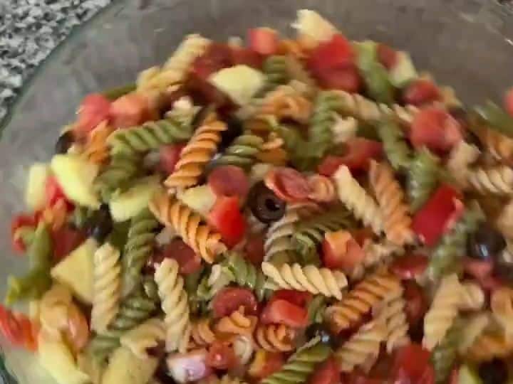 A large glass bowl filled with finished pasta salad showing pasta, tomatoes, cucumbers, pepperoni, and olives mixed together.