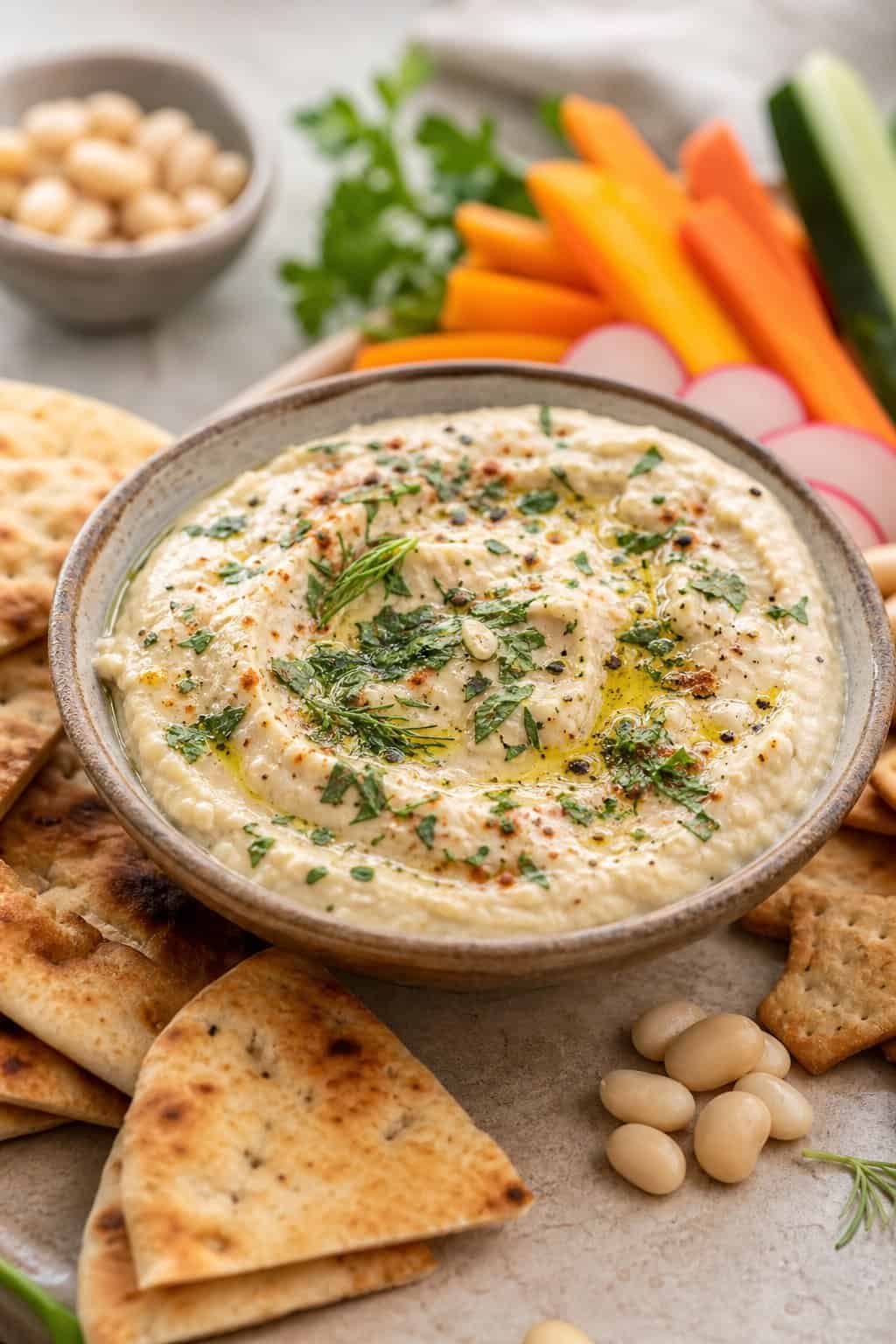 A bowl of creamy garlic-herb white bean dip surrounded by vegetables, pita wedges, and crackers.