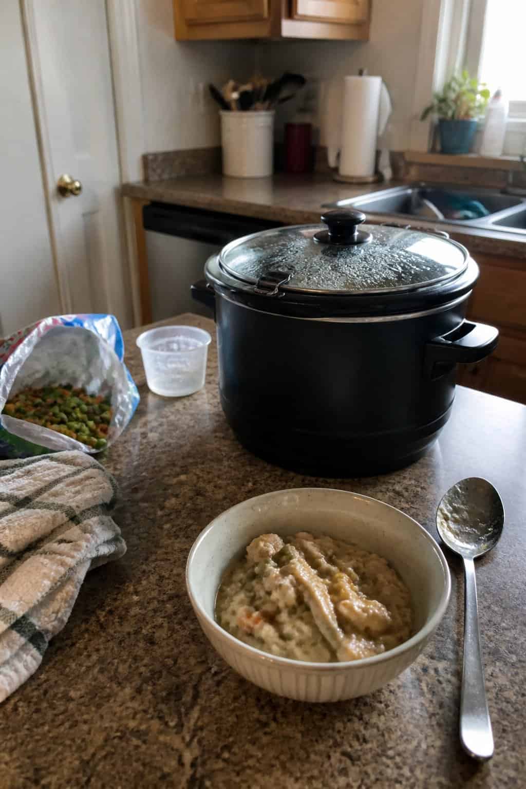 A portable slow cooker sits beside a bowl of a simple meal and everyday cooking tools on a kitchen countertop.