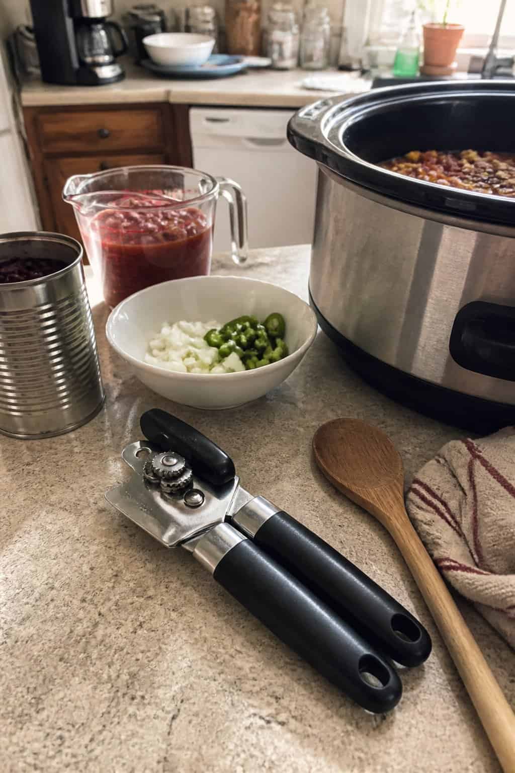 A manual can opener sits beside an open crockpot and ingredients on a kitchen counter.