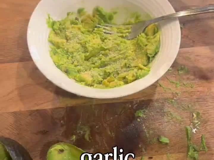 A bowl of mashed avocado guacamole is being stirred with a fork next to empty avocado shells on a wooden board.